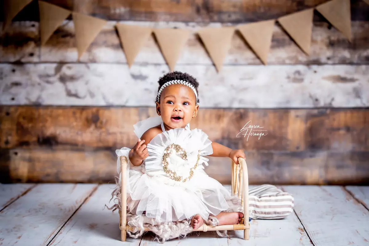 A baby girl sitting in a small wooden bed against a rustic wooden backdrop with paper bunting