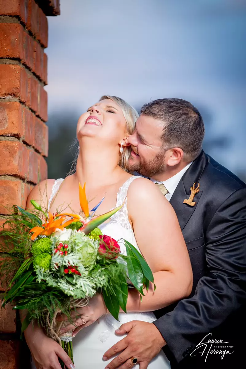 A bride laughing while holding a bouquet, and a groom smiling and nuzzling her neck