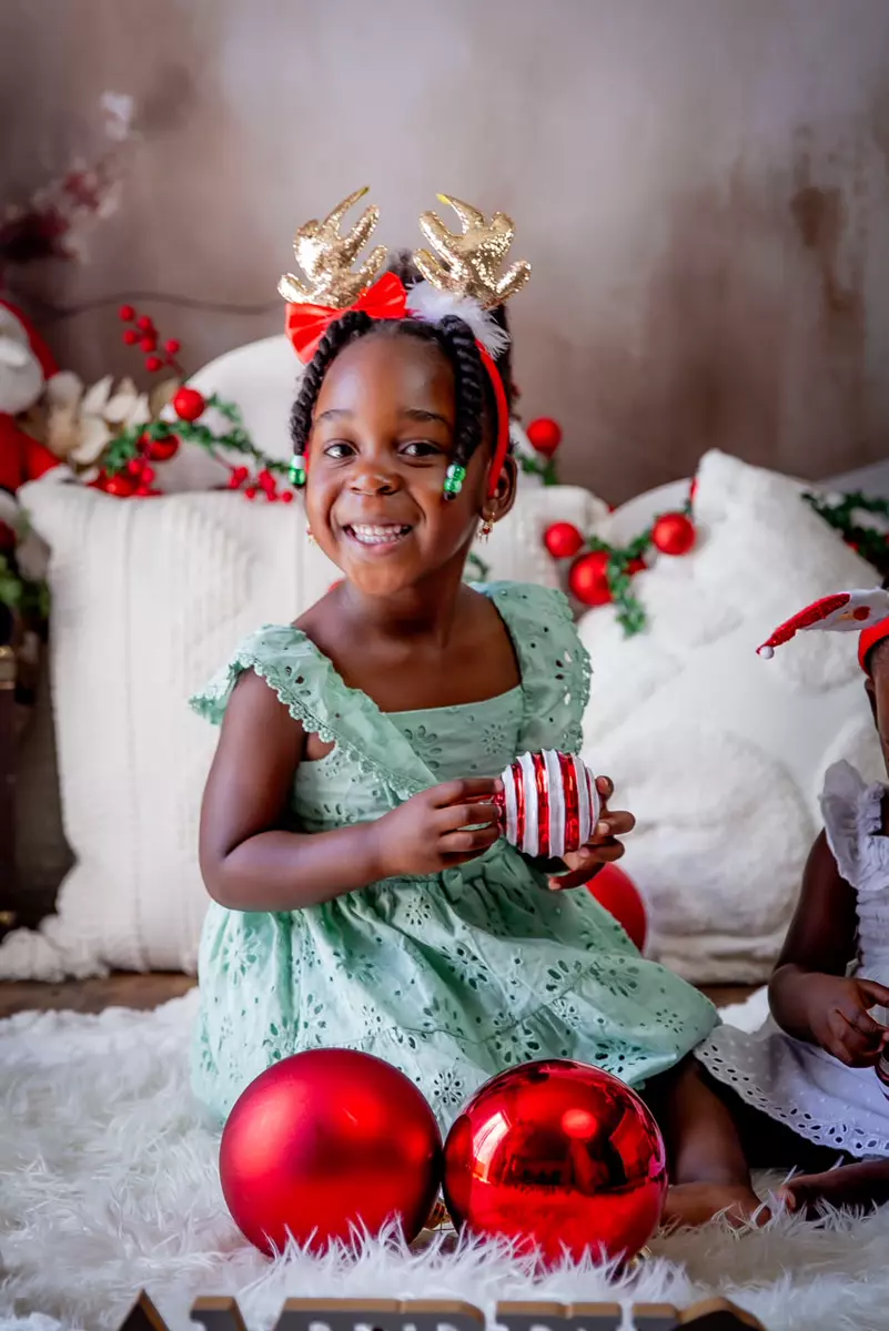 Young girl smiling, wearing reindeer antlers headband and a light green dress, holding a Christmas ornament