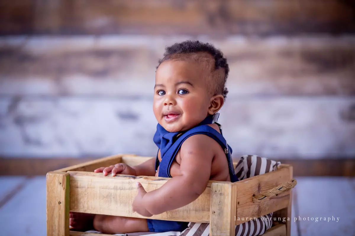 A cute baby sitting inside a wooden crate, wearing a dark blue outfit with suspenders and a bow tie