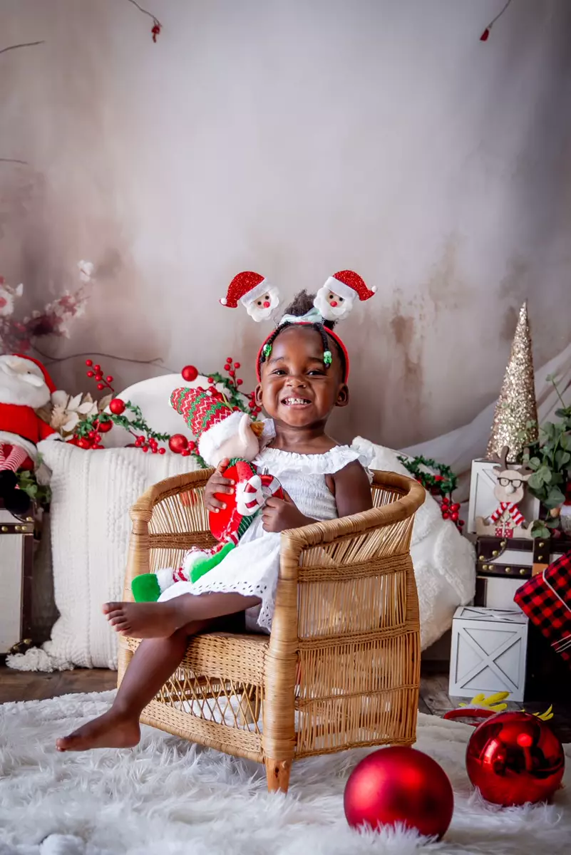 A smiling young child sitting in a wicker chair holding a Christmas toy, surrounded by holiday decorations