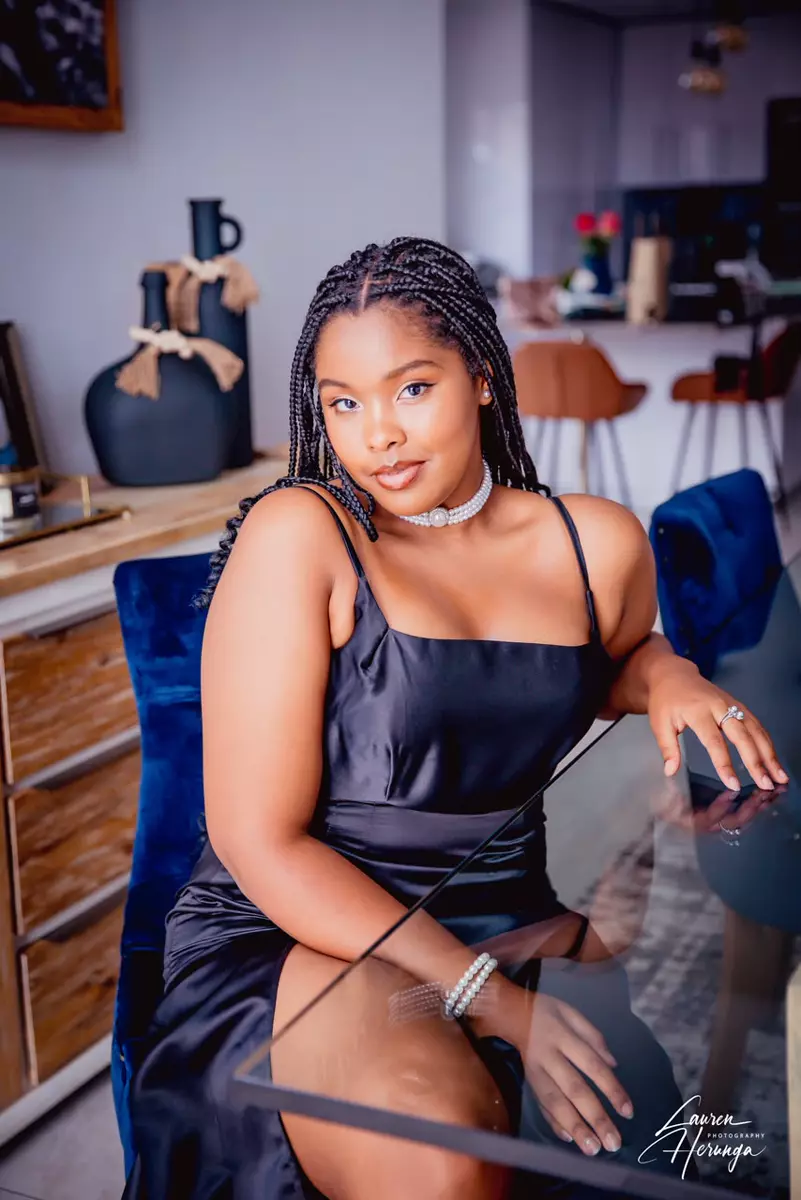 A young woman with braided hair wearing a black dress and pearl choker, seated at a glass table