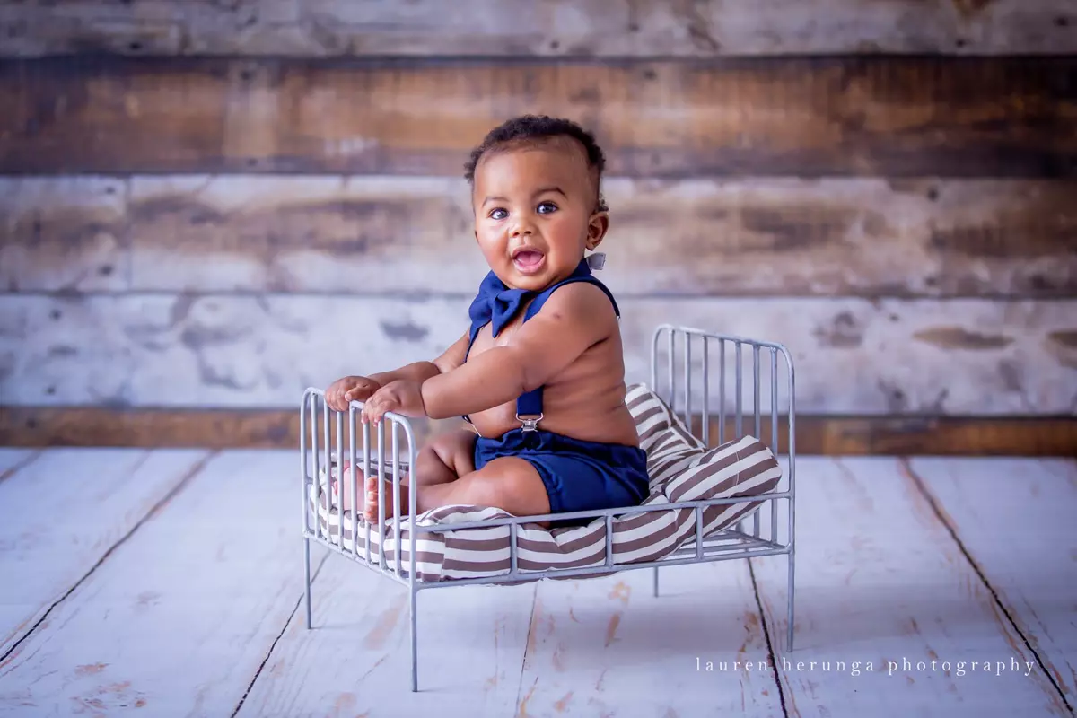 A baby boy sitting in a miniature metal bed wearing blue shorts and a bow tie
