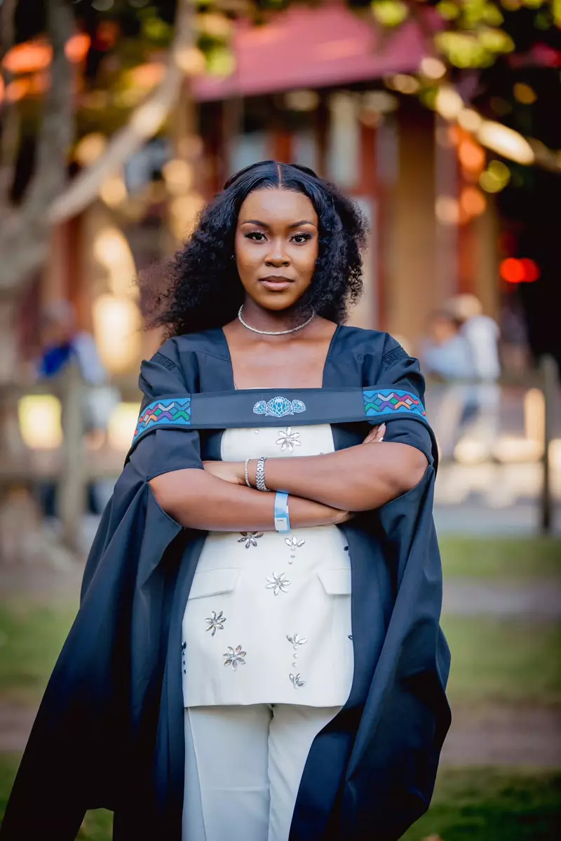 A woman standing outside, wearing a graduation gown over a white patterned outfit with arms crossed