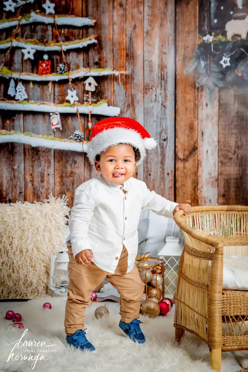 A toddler wearing a Santa hat and a white button-down shirt in front of a Christmas-themed backdrop