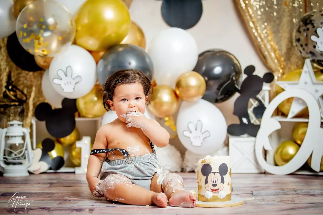 A toddler sitting on a floor with a cake, surrounded by Mickey Mouse-themed decorations
