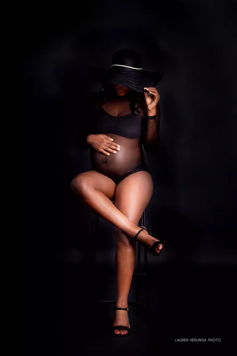 Pregnant woman in black outfit and hat sitting on a stool against a dark background