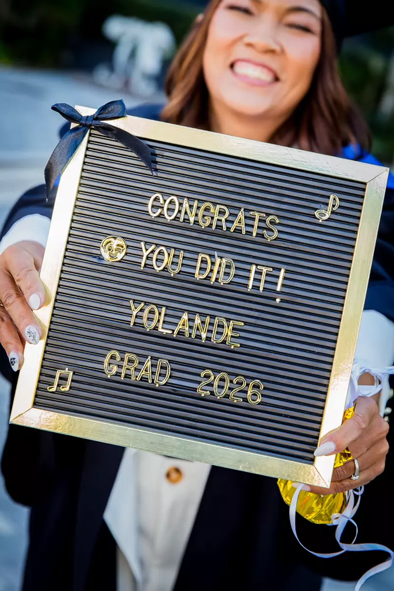 A smiling graduate holding a letter board that reads CONGRATS YOU DID IT