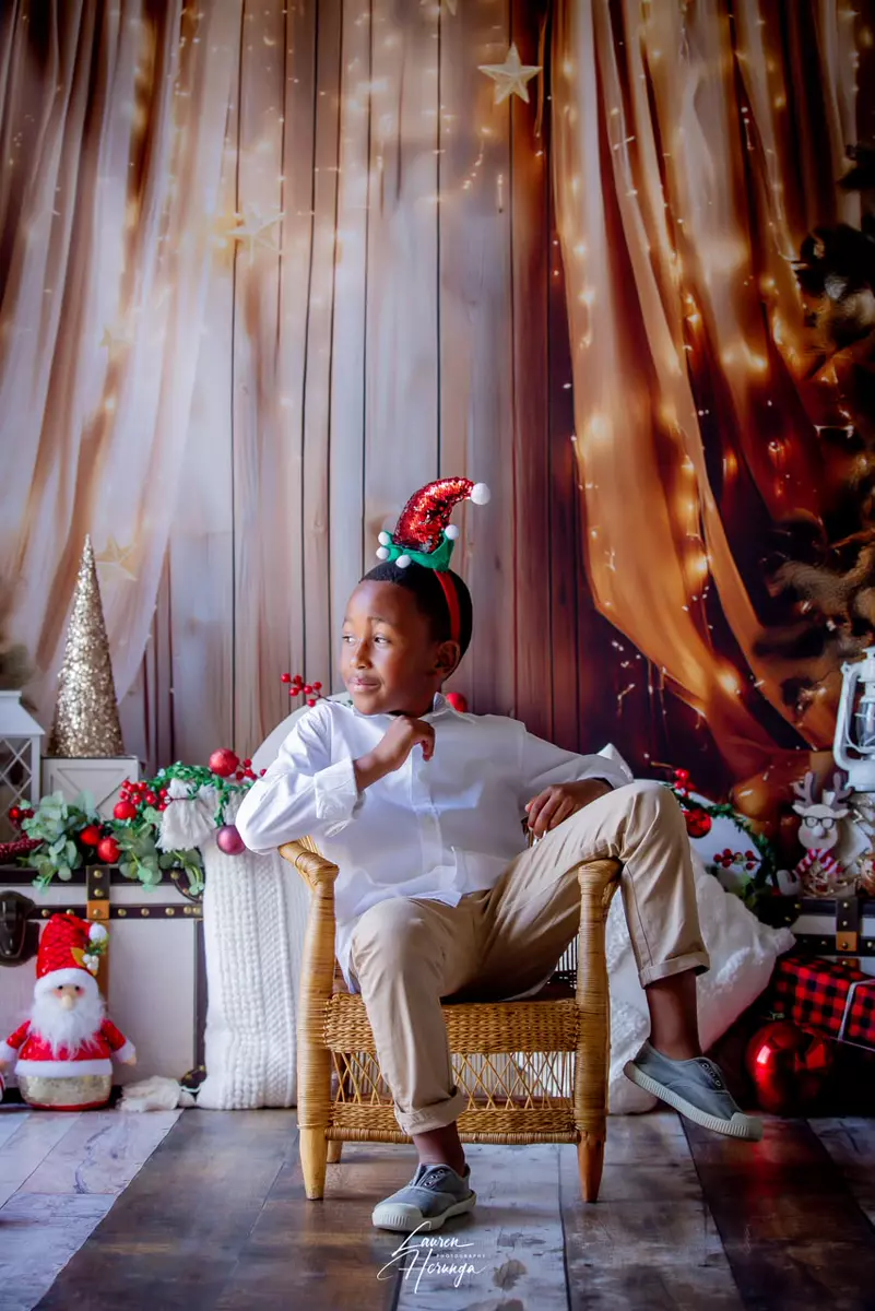 A young boy wearing a festive elf hat and seated in a chair against a Christmas-themed backdrop