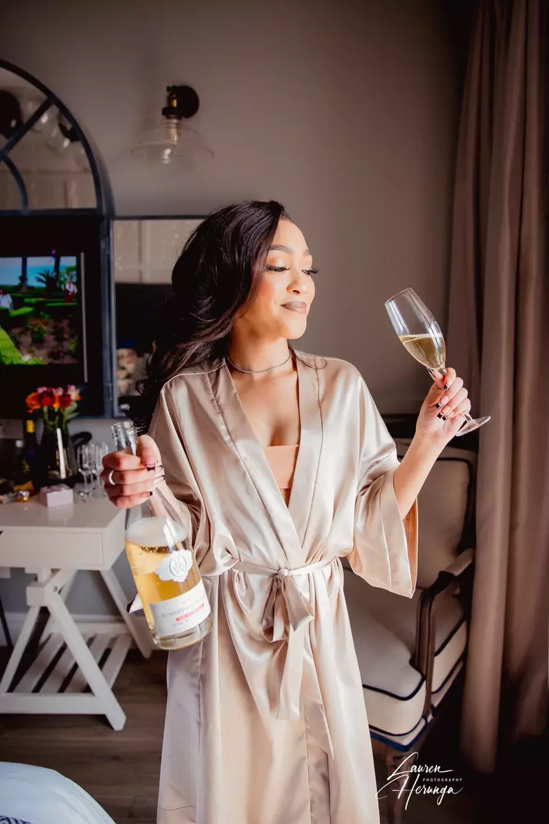 A woman in a satin robe holding a champagne bottle and glass in a bedroom